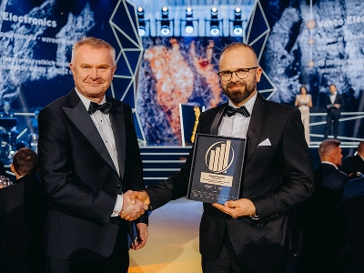 En bild på två män i formell smoking som skakar hand. Mannen till höger, Maciej Góralski, håller ett inramat pris med ett guldfärgat emblem som identifierar det som utmärkelsen Årets entreprenör 2022. I den suddiga bakgrunden syns en scen med abstrakta geometriska strukturer och dynamiska blå och rödorange grafiska mönster. Flera gäster sitter vid bord i den svagt upplysta evenemangshallen.