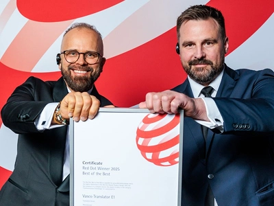 Två män i formell affärsklädsel – den vänstra i smoking med glasögon, den högra i mörkblå kostym – håller gemensamt ett inramat prisbevis med båda händerna. På certifikatet står det uttryckligen Red Dot Winner 2025 Best of the Best tilldelat Vasco Translator E1. I bakgrunden syns en storskalig, dynamisk grafisk bakgrund med abstrakta röda och vita vågmönster.
