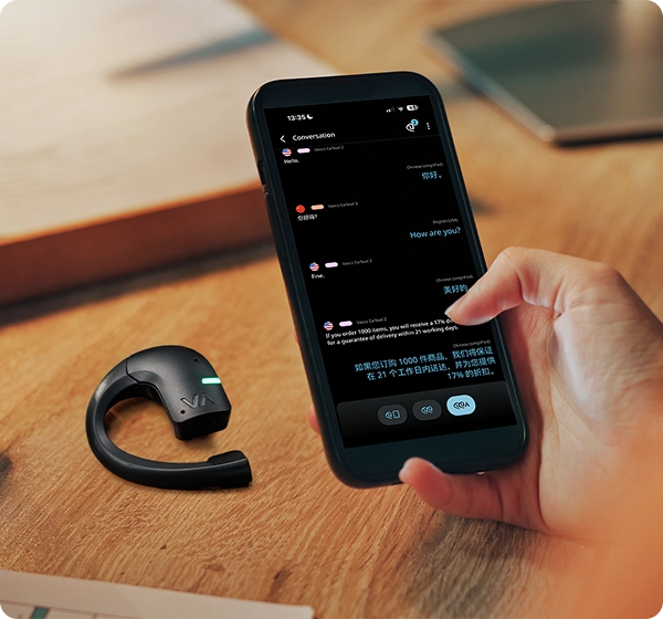 Närbild på en hand som håller en smartphone där en chatt med översättning i realtid mellan engelska och kinesiska visas. Bredvid telefonen ligger ett svart Vasco-översättningsöronsnäcka med en grön lysande LED-indikator på ett träbord. 