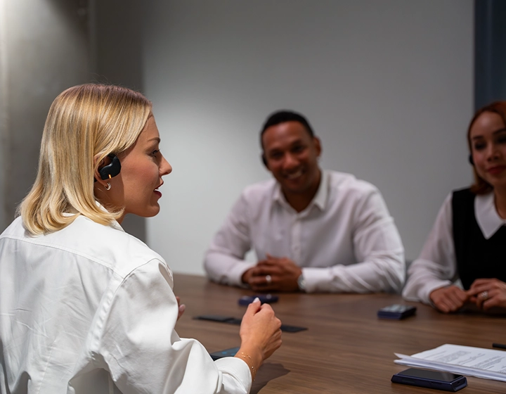 En blond kvinna i profil, klädd i en vit blus, talar under ett affärsmöte vid ett konferensbord av trä. Hon bär ett svart Vasco Translation E1-öronsnäcka. I den suddiga bakgrunden sitter två andra affärsmän på andra sidan bordet, leende och lyssnande uppmärksamt. 