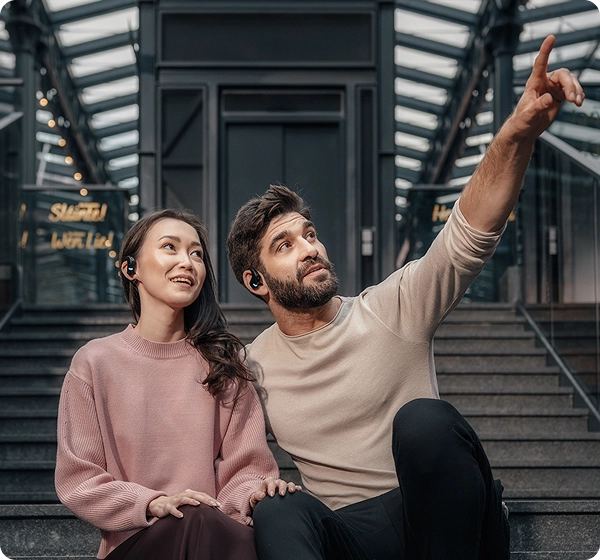 En man och en kvinna sitter på betongtrappsteg inuti en modern byggnad med ett glastak. Mannen pekar uppåt med handen, medan båda bär svarta Vasco Translation E1-hörlurar och tittar i den riktning han pekar. 
