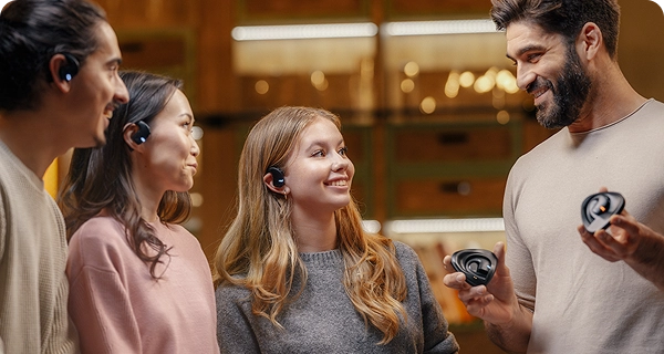 En grupp bestående av fyra personer med olika utseende visar upp den universella designen hos Vasco Translation E1-öronsnäckorna i ett rum med varm belysning. Tre av personerna bär de svarta öronsnäckorna och visar hur bekvämt de sitter i olika öronformer, medan en leende, skäggig man till höger håller i två laddningsfodral. 