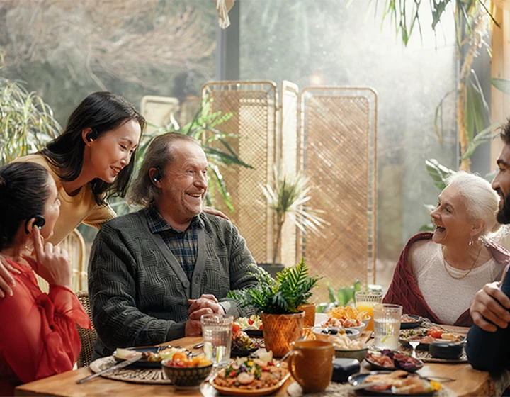En familj sitter runt ett bord med Vasco Translator E1-hörlurar, skrattar och pratar med varandra.