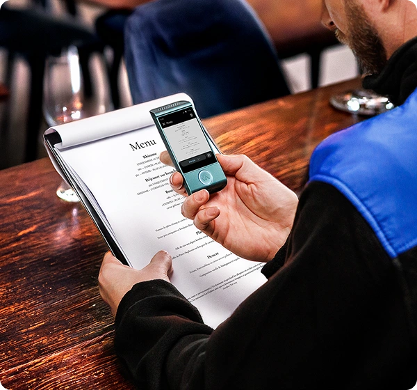 En närbild tagen uppifrån av en person som sitter vid ett träbord på en restaurang och använder en handhållen elektronisk översättningsenhet för att läsa menyn. Personen håller en vit pappersmeny i vänster hand samtidigt som hen med höger hand håller en liten, blågrön Vasco-översättare över den. Enhetens skärm visar en digital version av menyn som översätts till polska. Personen bär en svart jacka med en ljusblå axelpanel. Bakgrunden är mjukt suddig och visar den varma interiören i en restaurang. 