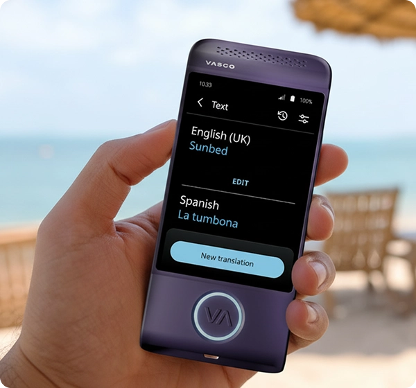 En närbild av en hand som håller en lila elektronisk översättare av märket Vasco. På skärmen visas en textöversättning från engelska (Storbritannien) till spanska, där ordet ”Sunbed” har översatts till ”La tumbona”. I bakgrunden syns en ljus, solig strand med en suddig vy av havet, gyllene sand, flera solstolar och ett strandparaply av strå. 
