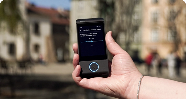 En närbild av en persons hand som håller en svart elektronisk översättare av märket Vasco, med en suddig utomhusbakgrund av ett torg med historiska byggnader. På enhetens skärm visas inställningarna för ”Översättningens läshastighet”, med ett skjutreglage inställt på ”Normal” och en anteckning som lyder: ”Justera översättningens läshastighet genom att flytta skjutreglaget.” Personen som håller i enheten bär ett silverarmband. Det starka dagsljuset belyser skärmens gränssnitt, vilket gör inställningarna lätta att se. 