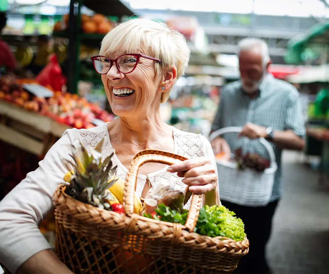 En leende äldre kvinna med kort blont hår och röda glasögon bär en flätad korg fylld med färsk frukt och grönsaker på en utomhusmarknad. I den suddiga bakgrunden handlar en annan person bland stånden med frukt och grönsaker.