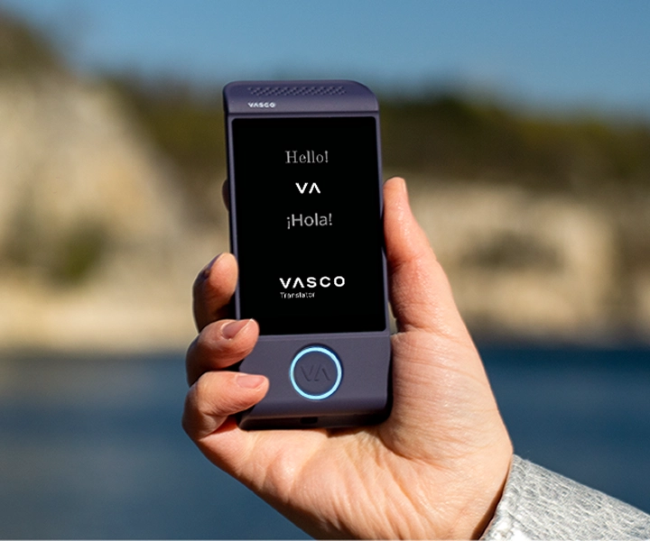 En närbild av en hand som håller en lila elektronisk översättningsenhet av märket Vasco Translator M4 mot en ljus utomhusbakgrund. På skärmen visas hälsningarna ”Hello!”, ”¡Hola!” och logotypen för ”VASCO Translator”.